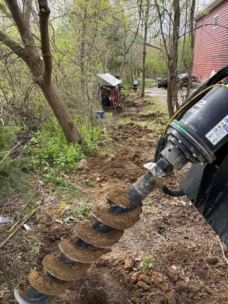 Close-up of auger bit drilling through soil in a wooded area for fence posthole installation